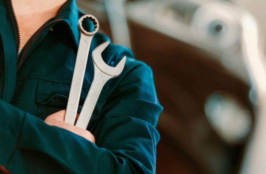 A person in a dark blue mechanic's uniform stands with arms crossed, holding two wrenches. One wrench has an open-end head, and the other has a box-end head. The background is blurred, showing the contours of a vehicle, suggesting an automotive repair setting at one of the top TESDA Accredited Competency Assessment Centers.