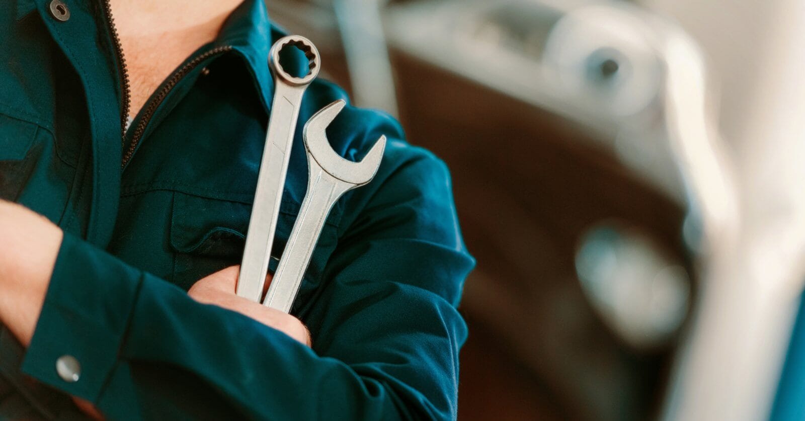 A person in a dark blue mechanic's uniform stands with arms crossed, holding two wrenches. One wrench has an open-end head, and the other has a box-end head. The background is blurred, showing the contours of a vehicle, suggesting an automotive repair setting at one of the top TESDA Accredited Competency Assessment Centers.