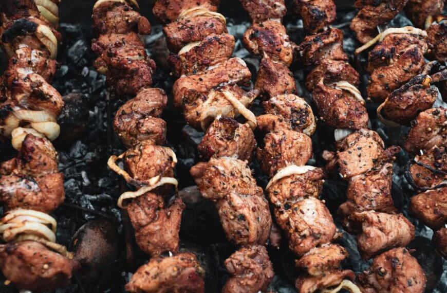 Several skewers of marinated meat and onions are being grilled over a bed of charcoal. The meat is slightly charred, giving it a deliciously cooked look with visible spices and seasoning. The skewers are spaced closely together, filling up the grill surface—just like you'd find at the best restaurants in Tomas Morato.