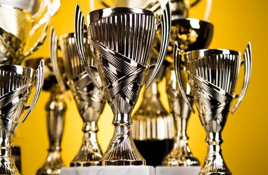 A collection of shiny silver trophies with intricate designs, featuring two handles each and mounted on marble bases, are displayed closely together. The background is a solid yellow color, emphasizing the polished surfaces and reflective qualities of these prestigious awards from top trophy suppliers.