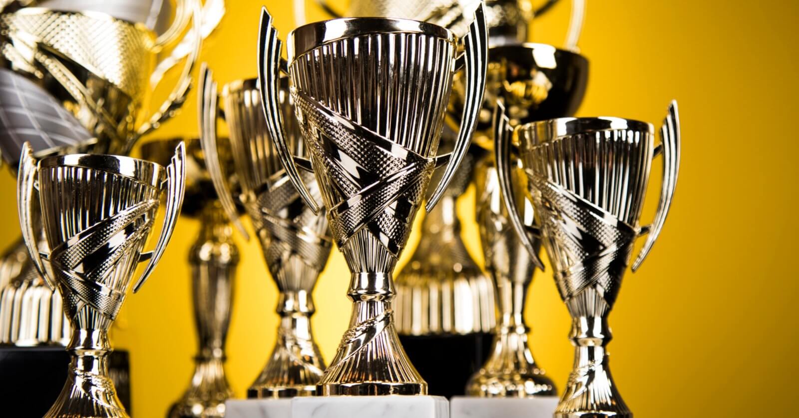 A collection of shiny silver trophies with intricate designs, featuring two handles each and mounted on marble bases, are displayed closely together. The background is a solid yellow color, emphasizing the polished surfaces and reflective qualities of these prestigious awards from top trophy suppliers.