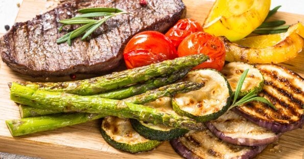 A grilled beef steak garnished with rosemary is served on a wooden board alongside grilled vegetables, including asparagus, zucchini slices, cherry tomatoes, eggplant slices, and bell pepper pieces. This fresh and appetizing meal embodies rustic elegance. Try it at one of the best restaurants in Vertis North.