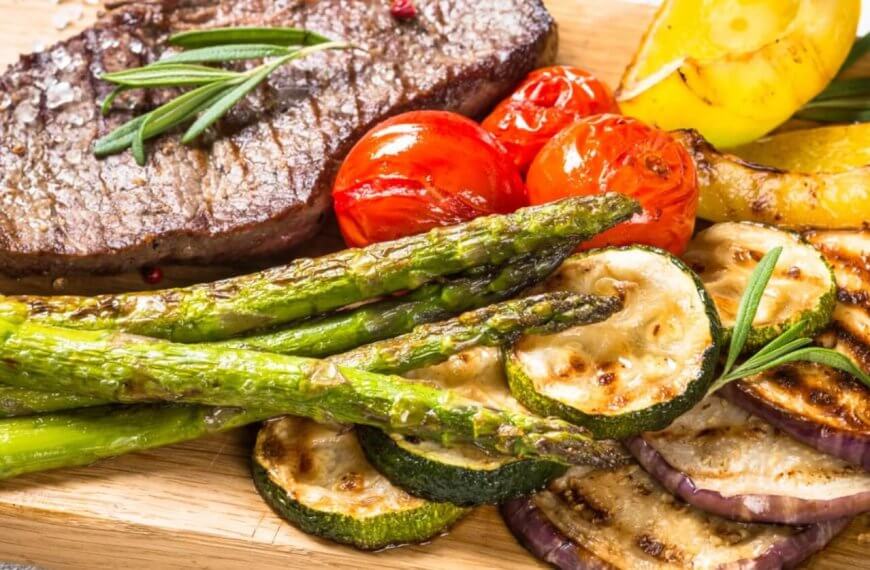 A grilled beef steak garnished with rosemary is served on a wooden board alongside grilled vegetables, including asparagus, zucchini slices, cherry tomatoes, eggplant slices, and bell pepper pieces. This fresh and appetizing meal embodies rustic elegance. Try it at one of the best restaurants in Vertis North.