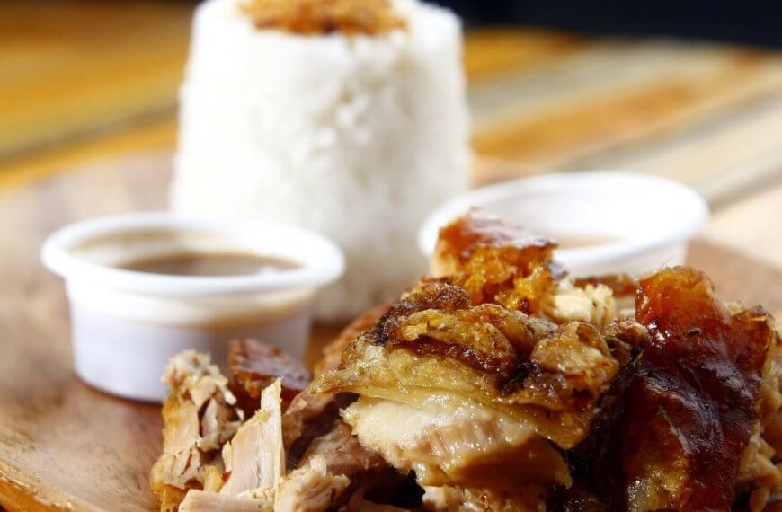 A wooden plate featuring a serving of lechon with crispy skin is shown in the foreground. Two small cups of dipping sauce are placed beside the pork. In the background, a mound of white rice topped with seasoning is visible, presenting a delightful foodie adventure at one of the must-try restaurants in Ayala Center Cebu.