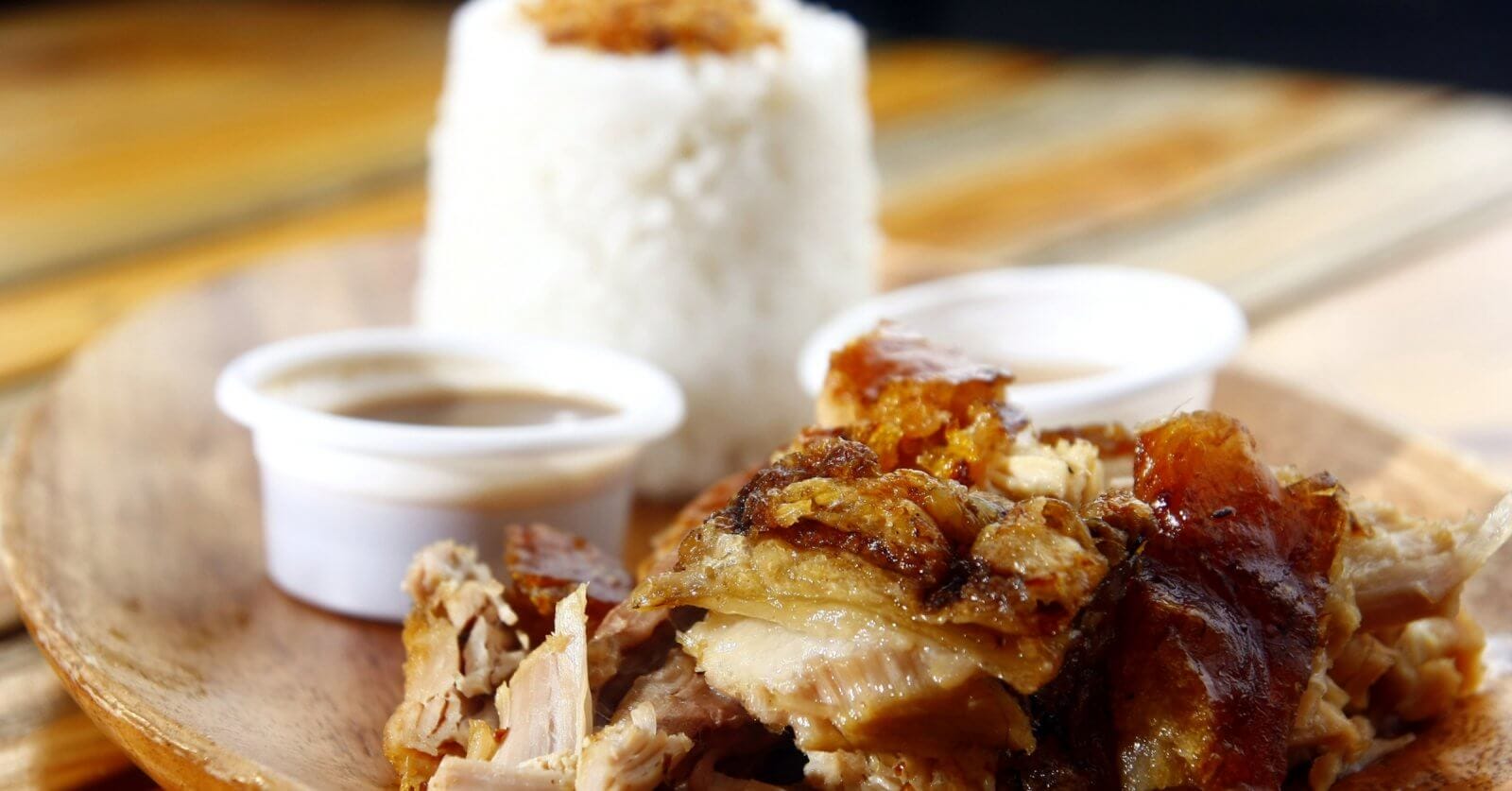 A wooden plate featuring a serving of lechon with crispy skin is shown in the foreground. Two small cups of dipping sauce are placed beside the pork. In the background, a mound of white rice topped with seasoning is visible, presenting a delightful foodie adventure at one of the must-try restaurants in Ayala Center Cebu.