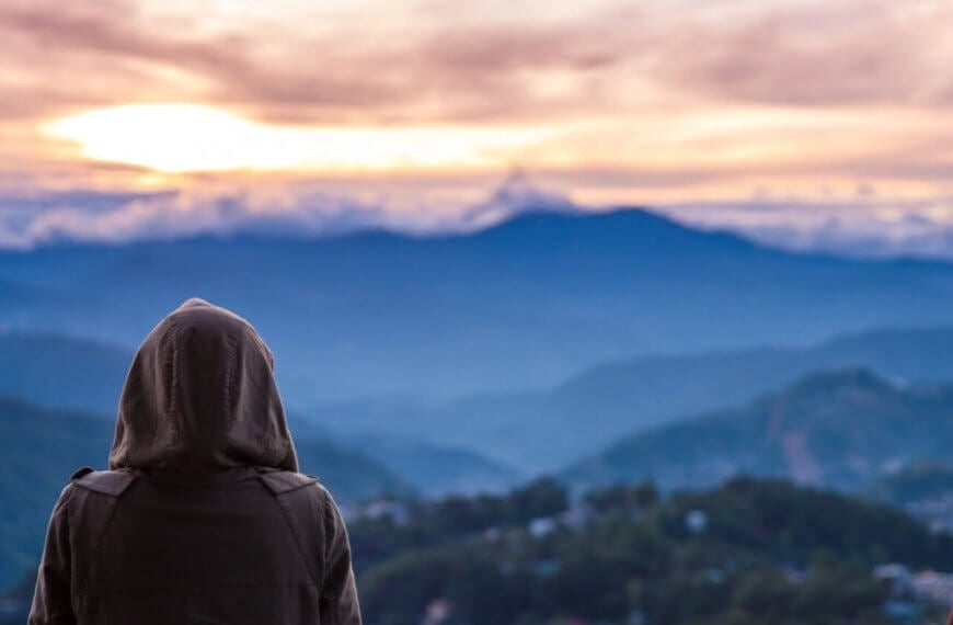 A person wearing a hooded jacket stands with their back to the camera, looking out over a vast, mountainous landscape at sunrise or sunset—a serene moment often experienced during a Baguio Tour. The sky is filled with soft clouds and warm light, casting a serene glow over the rolling hills and valleys below. A second person is partially visible.