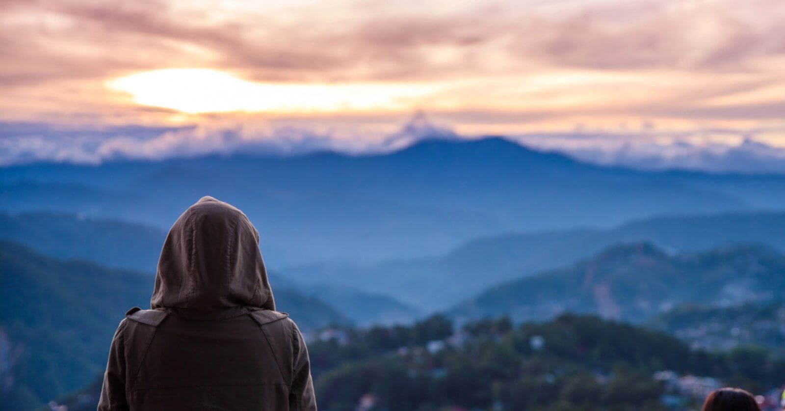 A person wearing a hooded jacket stands with their back to the camera, looking out over a vast, mountainous landscape at sunrise or sunset—a serene moment often experienced during a Baguio Tour. The sky is filled with soft clouds and warm light, casting a serene glow over the rolling hills and valleys below. A second person is partially visible.