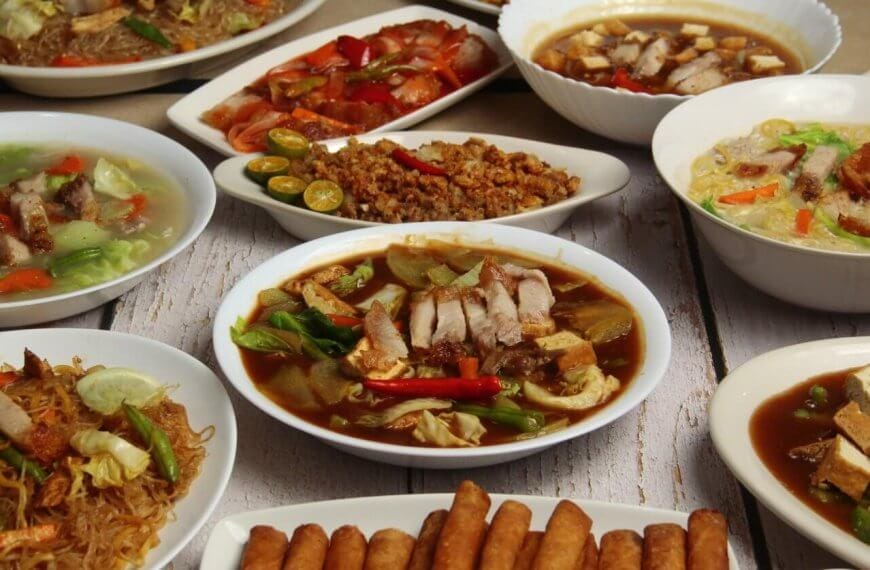 A spread of various Filipino dishes on white plates at one of Banawe, Quezon City's best restaurants. Dishes include bowls of noodle soup with vegetables and meat, a side of spring rolls, stir-fried noodles, grilled meat, and sliced vegetables. The dishes are richly colored with greens, reds, and browns on a white wooden table.