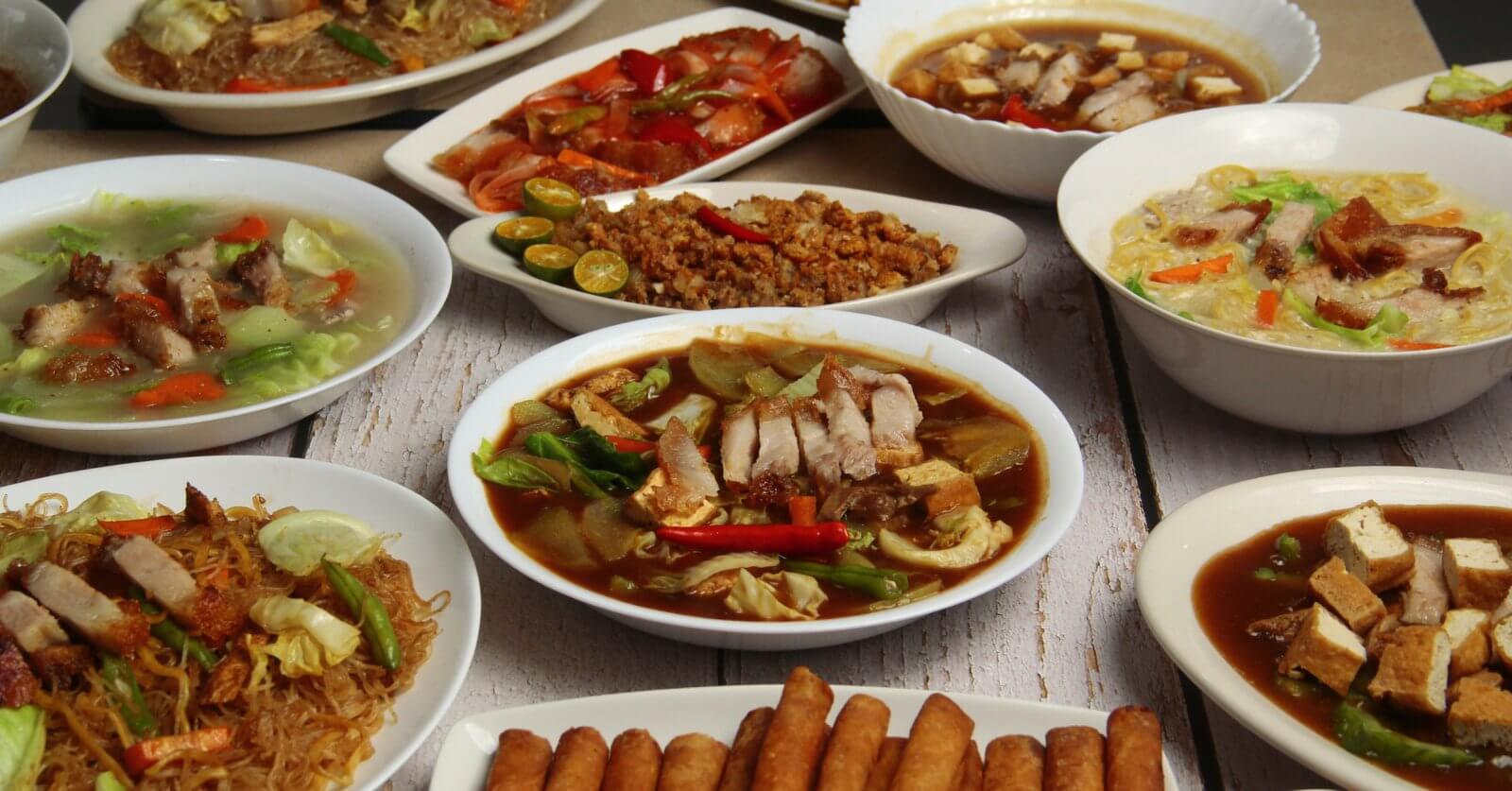 A spread of various Filipino dishes on white plates at one of Banawe, Quezon City's best restaurants. Dishes include bowls of noodle soup with vegetables and meat, a side of spring rolls, stir-fried noodles, grilled meat, and sliced vegetables. The dishes are richly colored with greens, reds, and browns on a white wooden table.