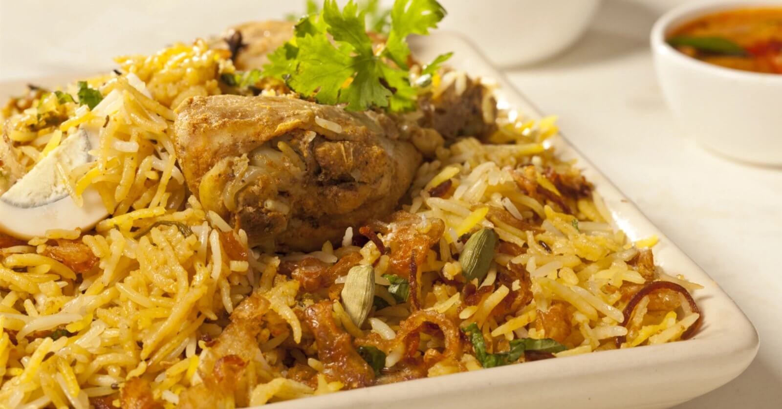 A close-up of a plate of biryani chicken featuring seasoned rice mixed with succulent pieces of chicken, garnished with fresh coriander leaves. This culinary experience is accompanied by a small bowl of curry sauce, slightly blurred in the background. The biryani is colorful, with shades of yellow and brown.
