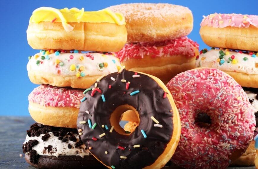 A vibrant assortment of donuts from the best donut shops in Metro Manila is stacked against a blue background. The donuts are topped with various icings and colorful sprinkles, including chocolate, pink, yellow, and white glazes. Some feature additional decorations like cookies and candy bits, making the scene playful and appetizing.