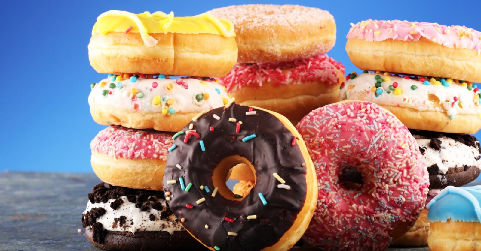 A vibrant assortment of donuts from the best donut shops in Metro Manila is stacked against a blue background. The donuts are topped with various icings and colorful sprinkles, including chocolate, pink, yellow, and white glazes. Some feature additional decorations like cookies and candy bits, making the scene playful and appetizing.