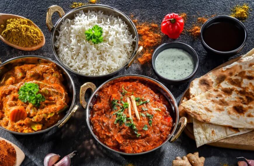 A colorful spread of Indian cuisine on a dark surface featuring bowls of white rice, chicken and vegetable curry, and a rich red curry garnished with cilantro and ginger. Accompanied by naan bread, mint yogurt sauce, and spices like curry powder, turmeric, and paprika—authentic flavors reminiscent of top Indian restaurants in Metro Manila.