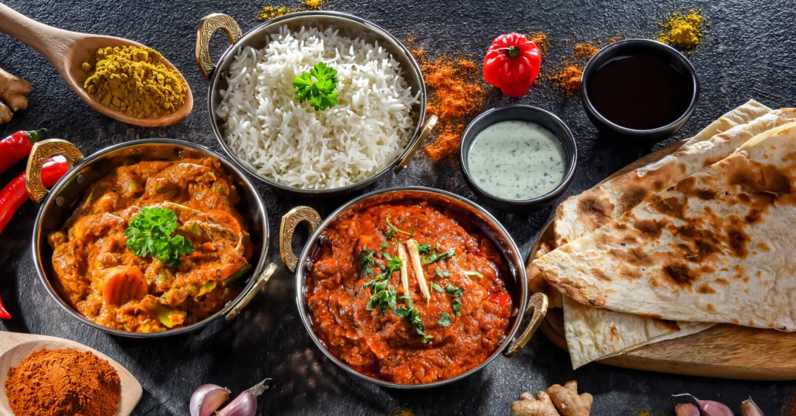 A colorful spread of Indian cuisine on a dark surface featuring bowls of white rice, chicken and vegetable curry, and a rich red curry garnished with cilantro and ginger. Accompanied by naan bread, mint yogurt sauce, and spices like curry powder, turmeric, and paprika—authentic flavors reminiscent of top Indian restaurants in Metro Manila.