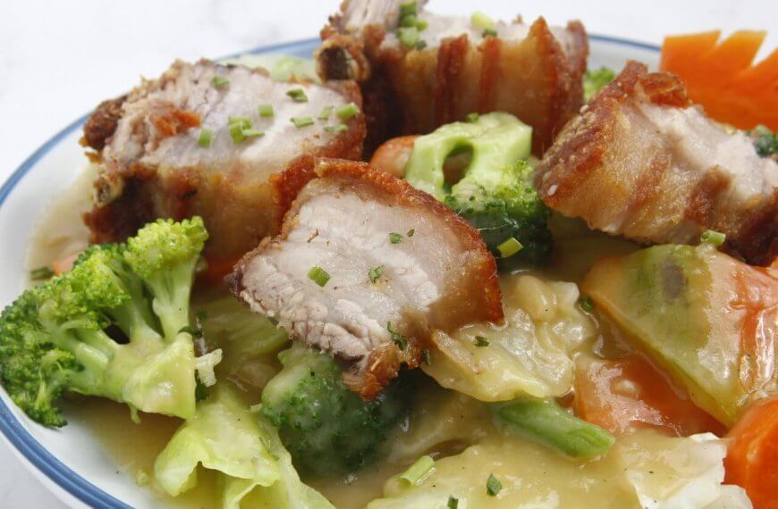A bowl of crispy pork belly pieces with a golden-brown exterior, served on a bed of vegetables including broccoli, carrots, and cabbage, all coated in a creamy, savory sauce. This dish from one of Nuvali's best restaurants is garnished with finely chopped chives, adding a touch of green.