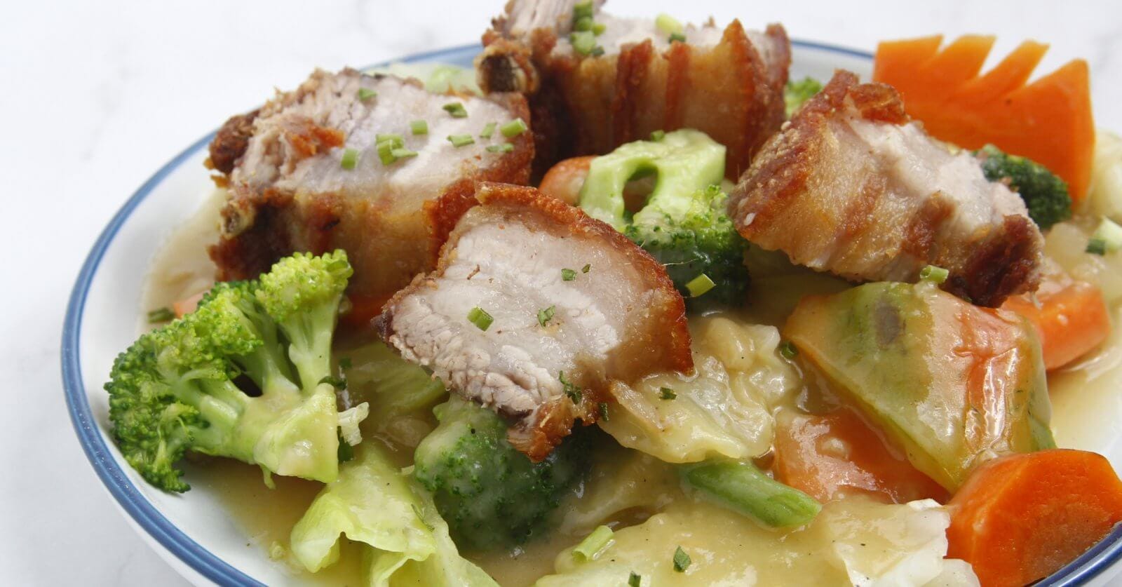 A bowl of crispy pork belly pieces with a golden-brown exterior, served on a bed of vegetables including broccoli, carrots, and cabbage, all coated in a creamy, savory sauce. This dish from one of Nuvali's best restaurants is garnished with finely chopped chives, adding a touch of green.
