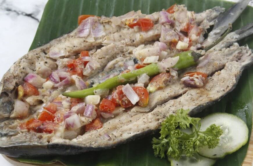 A grilled Bangus split open and laid flat on a green leaf, topped with chopped tomatoes, onions, and garlic, and garnished with a green chili pepper and a small bunch of parsley. Next to the fish are cucumber slices, all arranged on a woven mat—a classic dish you might find at 44 Must-Try Restaurants in Market! Market!.
