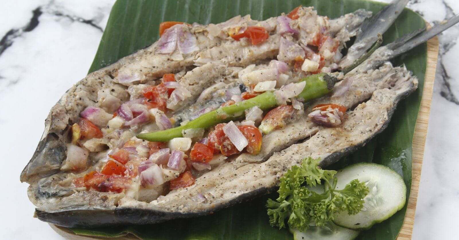 A grilled Bangus split open and laid flat on a green leaf, topped with chopped tomatoes, onions, and garlic, and garnished with a green chili pepper and a small bunch of parsley. Next to the fish are cucumber slices, all arranged on a woven mat—a classic dish you might find at 44 Must-Try Restaurants in Market! Market!.