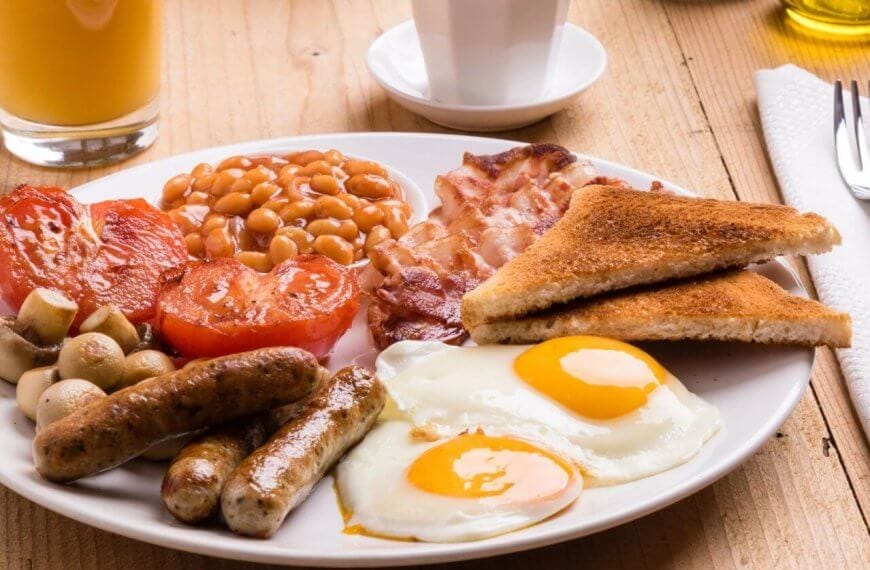 A plate of English breakfast featuring two sunny-side-up eggs, grilled tomatoes, baked beans, crispy bacon, sausages, grilled mushrooms, and two slices of toasted bread. Beside the plate are a fork and knife on a napkin, a glass of orange juice, and a white cup on a wooden table in one of BGC’s best breakfast restaurants.