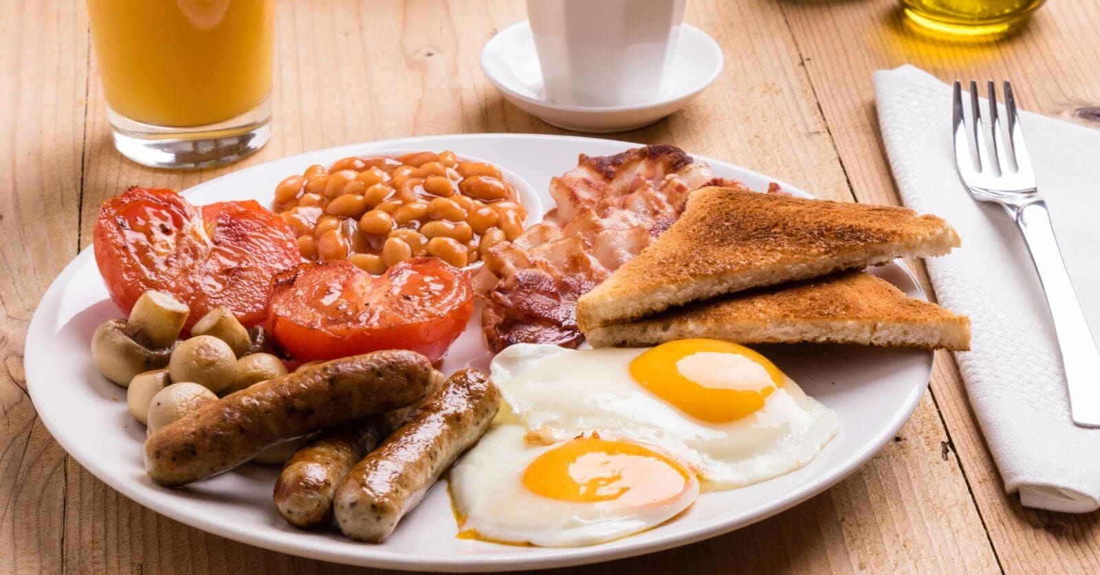 A plate of English breakfast featuring two sunny-side-up eggs, grilled tomatoes, baked beans, crispy bacon, sausages, grilled mushrooms, and two slices of toasted bread. Beside the plate are a fork and knife on a napkin, a glass of orange juice, and a white cup on a wooden table in one of BGC’s best breakfast restaurants.