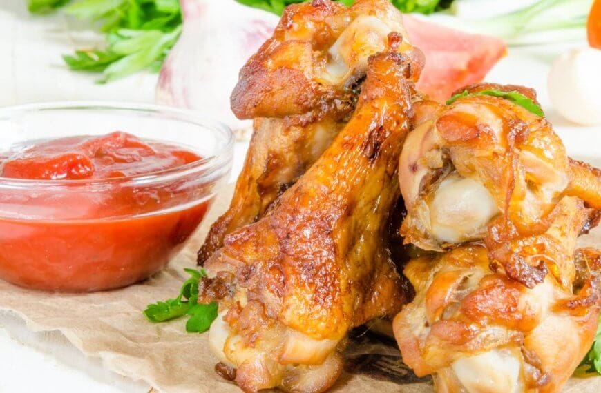 A close-up of golden-brown, crispy Korean chicken wings stacked on a rustic surface, garnished with fresh parsley. A small, clear bowl of red ketchup is positioned to the left. In the blurred background, there are assorted vegetables, including green herbs and garlic cloves—a scene reminiscent of Metro Manila's popular beer spots.