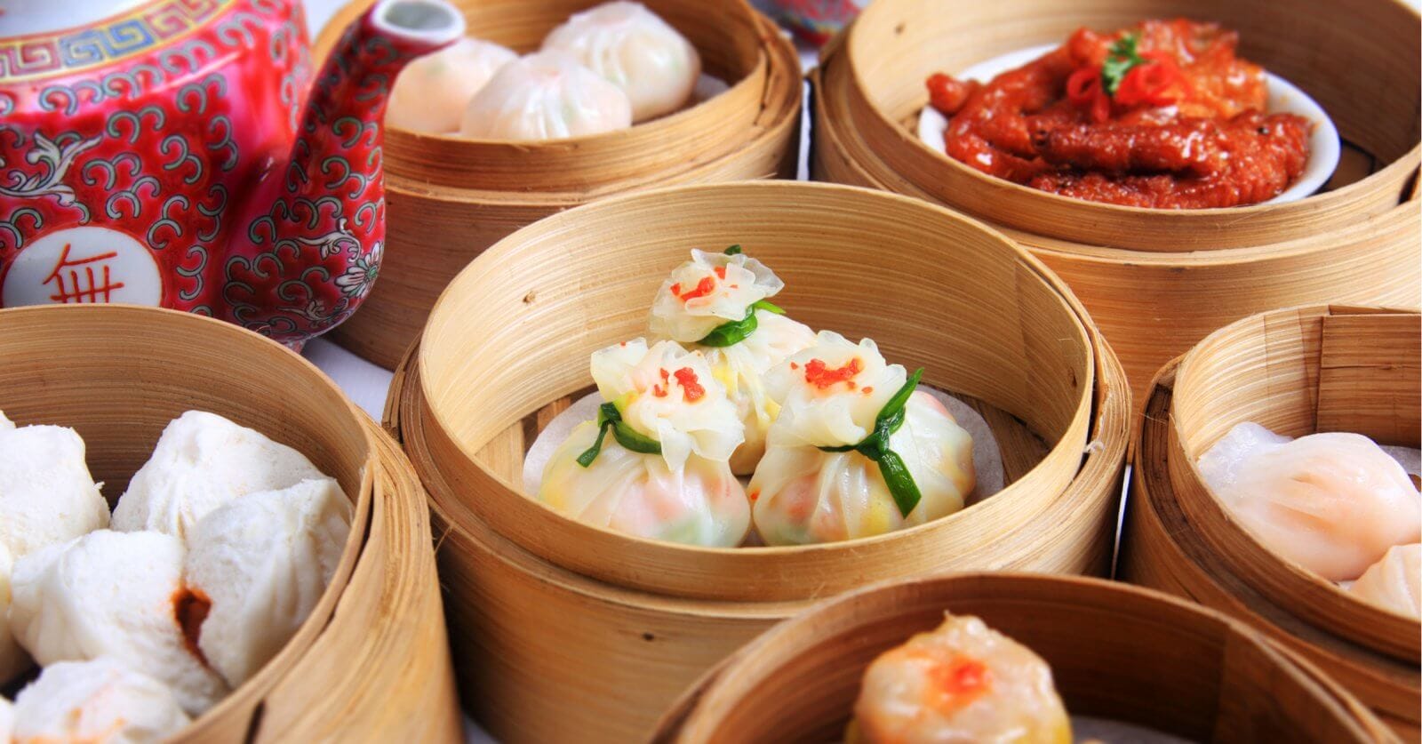 A variety of dimsum dishes are served in bamboo steamers. The dishes include dumplings, buns, and chicken feet. The dim sum items are intricately plated, with some garnished with a touch of red. A red, patterned teapot with Chinese characters is visible in the background—a typical scene in top 110 Chinese restaurants in Metro Manila.