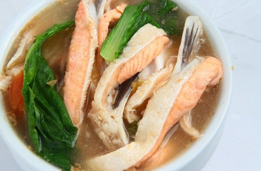 A bowl of clear soup contains pieces of fish, vibrant green leafy vegetables, and orange carrot slices. The fish appears to be salmon, with its distinctive pink color. The broth is light and clear, beautifully presenting a sulit dining experience that could rival the 40 best restaurants in Circuit Manila.