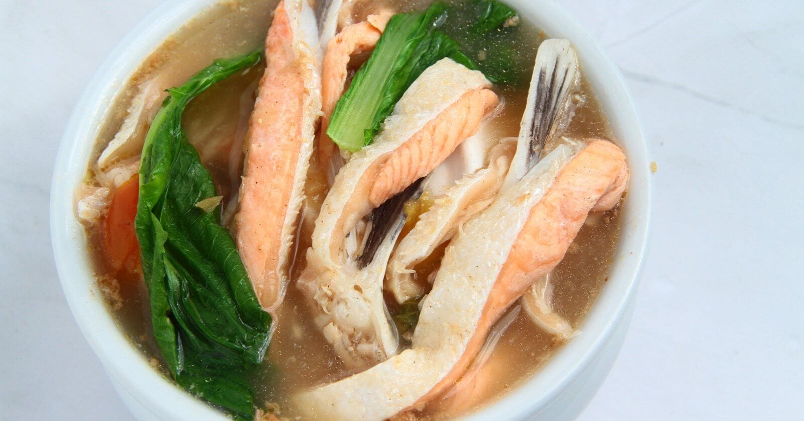 A bowl of clear soup contains pieces of fish, vibrant green leafy vegetables, and orange carrot slices. The fish appears to be salmon, with its distinctive pink color. The broth is light and clear, beautifully presenting a sulit dining experience that could rival the 40 best restaurants in Circuit Manila.
