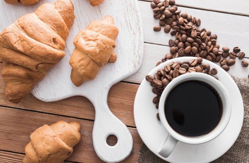 A white cup filled with black coffee from Sulit Coffee sits on a saucer with coffee beans scattered around it on a rustic wooden table. Next to the coffee, there is a wooden cutting board with golden brown croissants placed on it, and more croissants are scattered around the table, evoking the charm of a Metro Manila cafe bakery.
