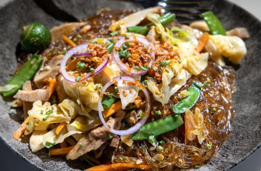 A close-up of a plate of Filipino pancit, featuring glass noodles mixed with sautéed vegetables like cabbage, carrot slices, and snow peas, topped with thinly sliced red onions, fried garlic bits, and chopped green onions. Often found in the best restaurants around Cubao, a green calamansi is seen on the side for garnish.