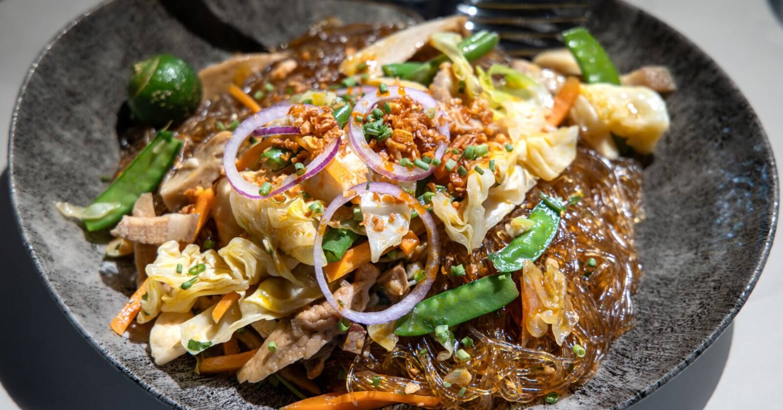 A close-up of a plate of Filipino pancit, featuring glass noodles mixed with sautéed vegetables like cabbage, carrot slices, and snow peas, topped with thinly sliced red onions, fried garlic bits, and chopped green onions. Often found in the best restaurants around Cubao, a green calamansi is seen on the side for garnish.