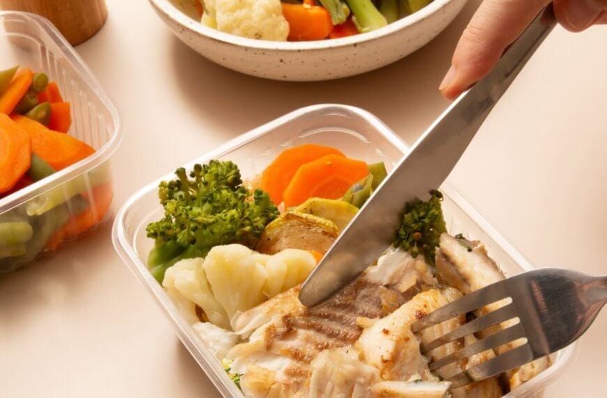 A person using a knife and fork to cut pieces of grilled fish in a plastic container. The container also holds cooked broccoli, cauliflower, and carrots. Additional containers filled with more steamed mixed vegetables and a white bowl are in the background, ideal for healthy meal plans from diet delivery services.