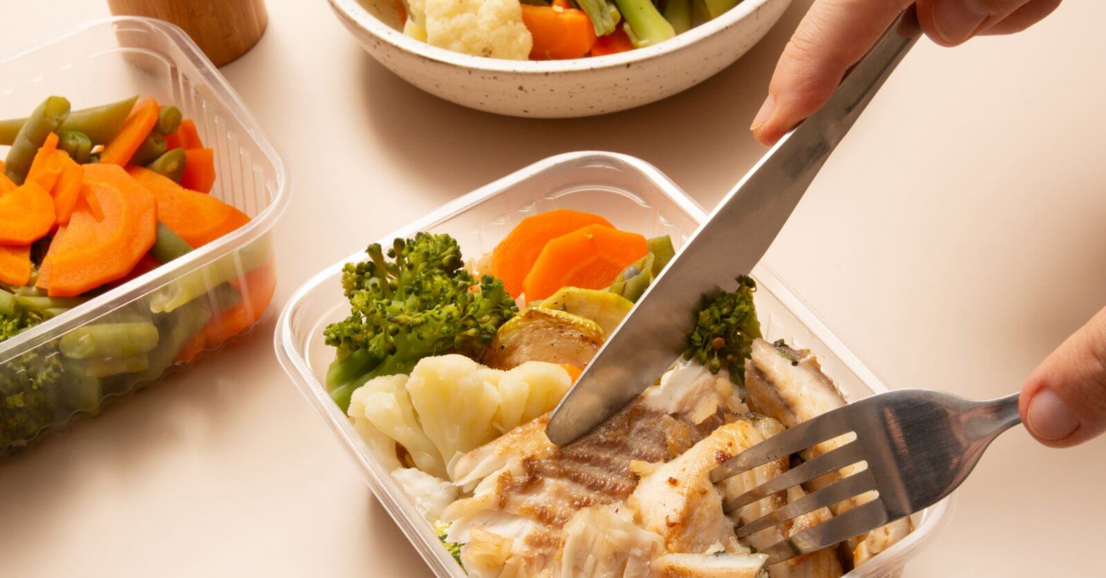 A person using a knife and fork to cut pieces of grilled fish in a plastic container. The container also holds cooked broccoli, cauliflower, and carrots. Additional containers filled with more steamed mixed vegetables and a white bowl are in the background, ideal for healthy meal plans from diet delivery services.