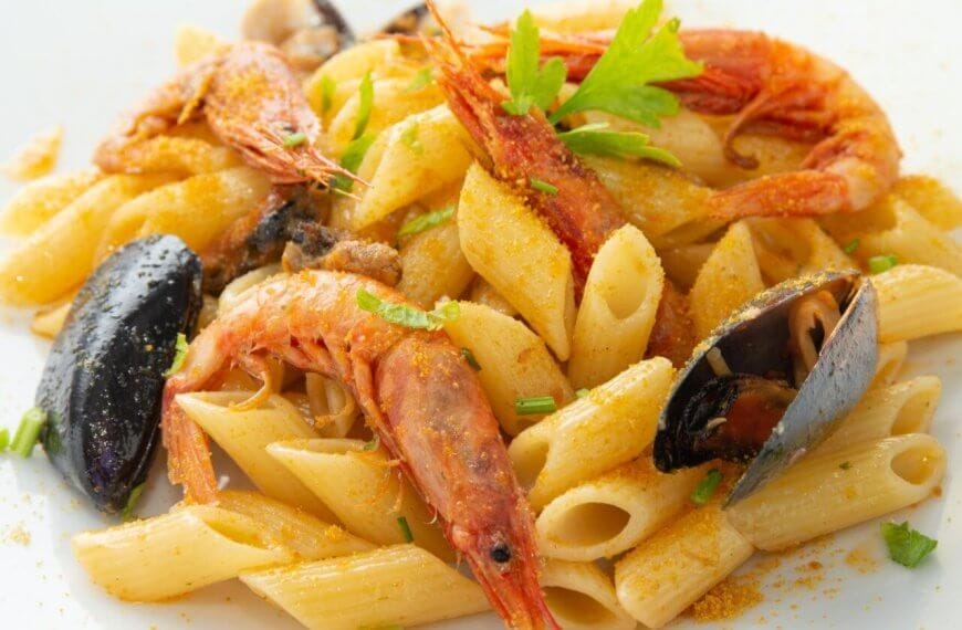A close-up of a seafood pasta dish featuring penne pasta, large shrimp, and mussels. Garnished with sprigs of fresh parsley and a light dusting of seasoning, the dish is served on a square white plate at one of the best restaurants in Evia Lifestyle Center. The vibrant colors contrast with the creamy pasta.