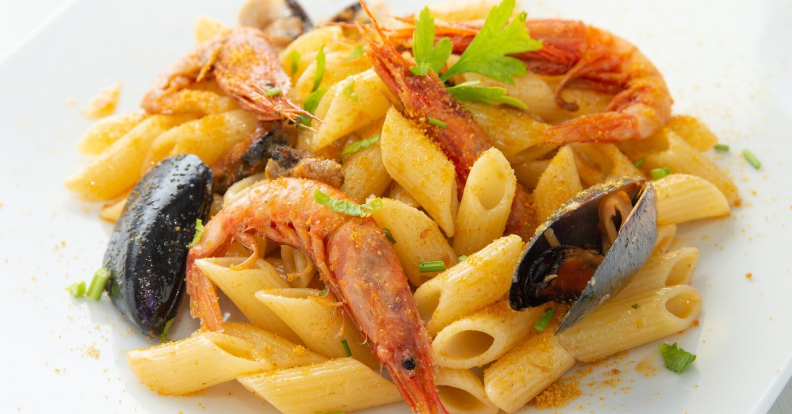 A close-up of a seafood pasta dish featuring penne pasta, large shrimp, and mussels. Garnished with sprigs of fresh parsley and a light dusting of seasoning, the dish is served on a square white plate at one of the best restaurants in Evia Lifestyle Center. The vibrant colors contrast with the creamy pasta.