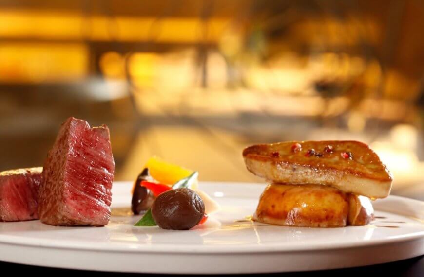 A gourmet dish on a white plate features a neatly cut piece of medium-rare beef, an artistic arrangement of colorful vegetables, a dollop of dark sauce, and seared foie gras atop a round base. This fine dining experience in Metro Manila is beautifully highlighted against a softly blurred background.