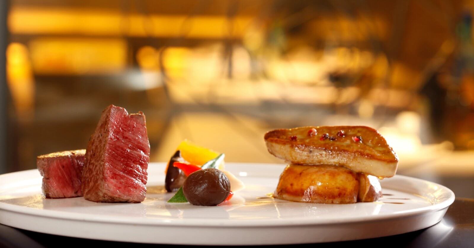 A gourmet dish on a white plate features a neatly cut piece of medium-rare beef, an artistic arrangement of colorful vegetables, a dollop of dark sauce, and seared foie gras atop a round base. This fine dining experience in Metro Manila is beautifully highlighted against a softly blurred background.