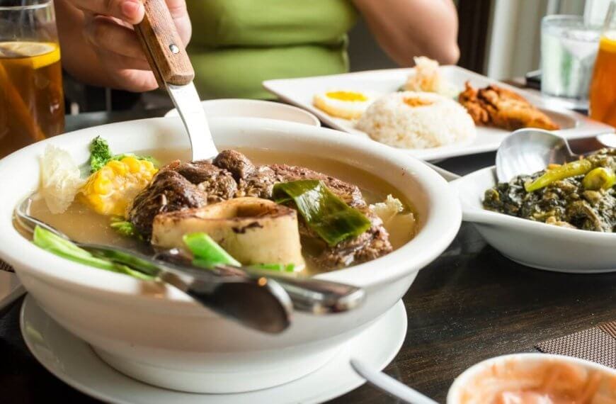 A person in a green shirt is cutting meat in a bowl of soup with a knife and fork. The table is set with iced tea, a plate of rice with fried food, and a dish of greens, highlighting the Best Restaurants. Small plates around feature a variety of foods, making it the Ultimate Glorietta Experience.