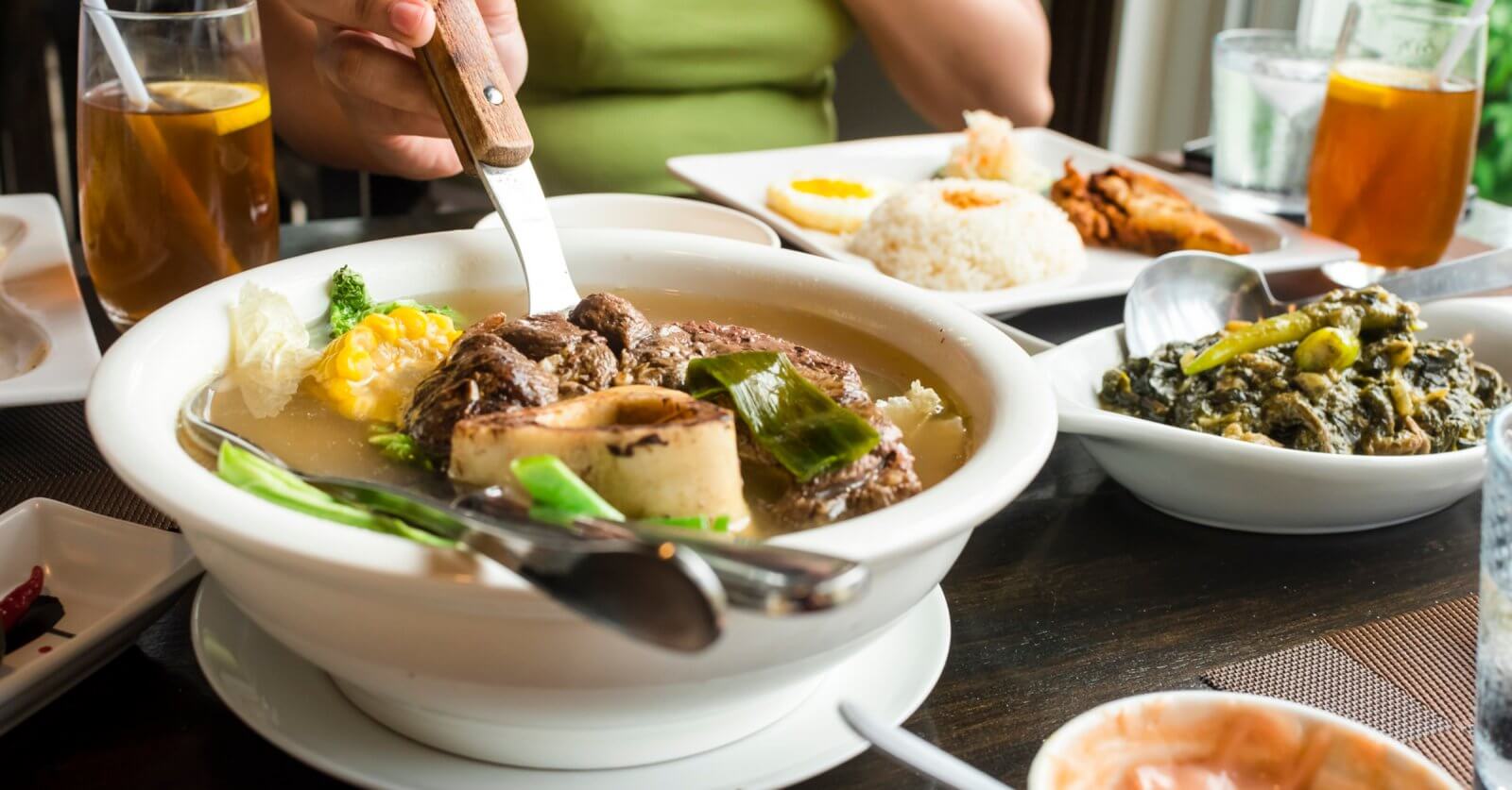 A person in a green shirt is cutting meat in a bowl of soup with a knife and fork. The table is set with iced tea, a plate of rice with fried food, and a dish of greens, highlighting the Best Restaurants. Small plates around feature a variety of foods, making it the Ultimate Glorietta Experience.