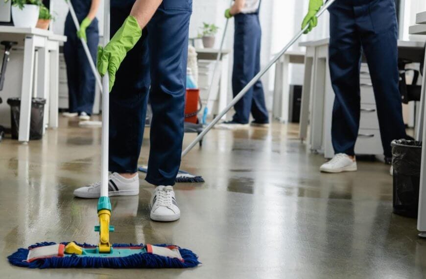 Cleaning people in blue uniforms and green gloves are mopping the floor in an office setting, representing Sulit Clean's Home Cleaning Services. The focus is on the person in the foreground, visible from the waist down, using a flat mop. White desks, office chairs, and trash cans fill out the Metro Manila background.