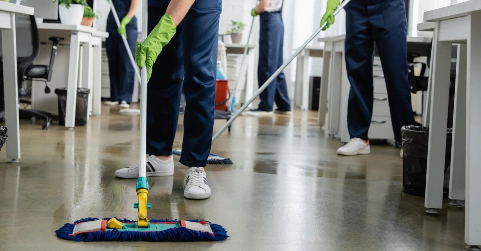 Cleaning people in blue uniforms and green gloves are mopping the floor in an office setting, representing Sulit Clean's Home Cleaning Services. The focus is on the person in the foreground, visible from the waist down, using a flat mop. White desks, office chairs, and trash cans fill out the Metro Manila background.