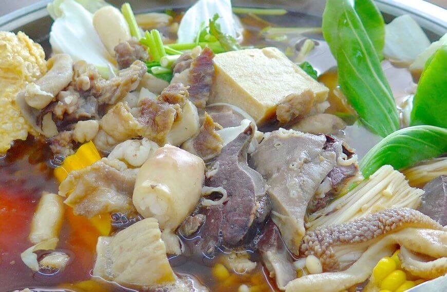An assortment of ingredients simmering in a hot pot filled with broth. Visible items include various cuts of meat, tofu, mushrooms, leafy greens, pieces of corn, and a chunk of tomato. The ingredients float in a rich brown broth, creating a hearty, communal meal—perfect for those looking to spice up their taste buds at the best hotpot restaurants in Metro Manila.