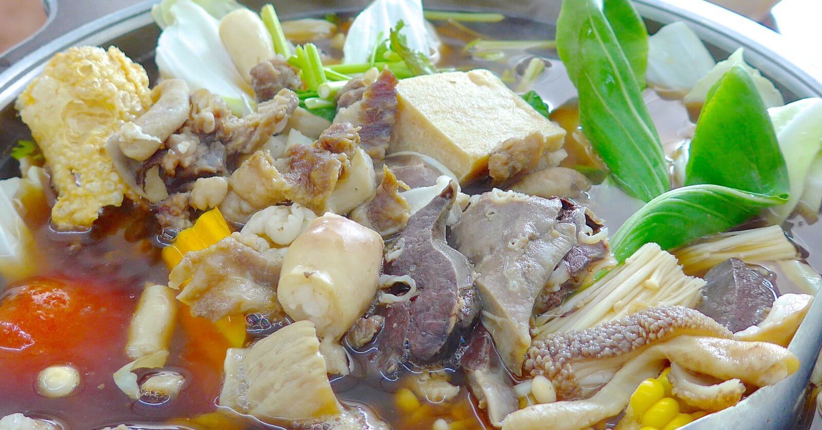 An assortment of ingredients simmering in a hot pot filled with broth. Visible items include various cuts of meat, tofu, mushrooms, leafy greens, pieces of corn, and a chunk of tomato. The ingredients float in a rich brown broth, creating a hearty, communal meal—perfect for those looking to spice up their taste buds at the best hotpot restaurants in Metro Manila.
