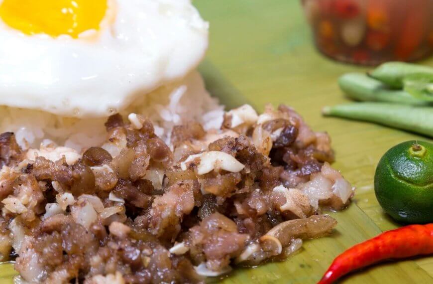 A close-up of traditional Filipino food featuring a serving of garlic rice topped with a sunny-side-up egg. Adjacent to the rice is a portion of sizzling chopped meat, likely pork. Garnishing the plate are a whole calamansi, a red chili pepper, and a few green beans. A condiment jar is blurred in the background.