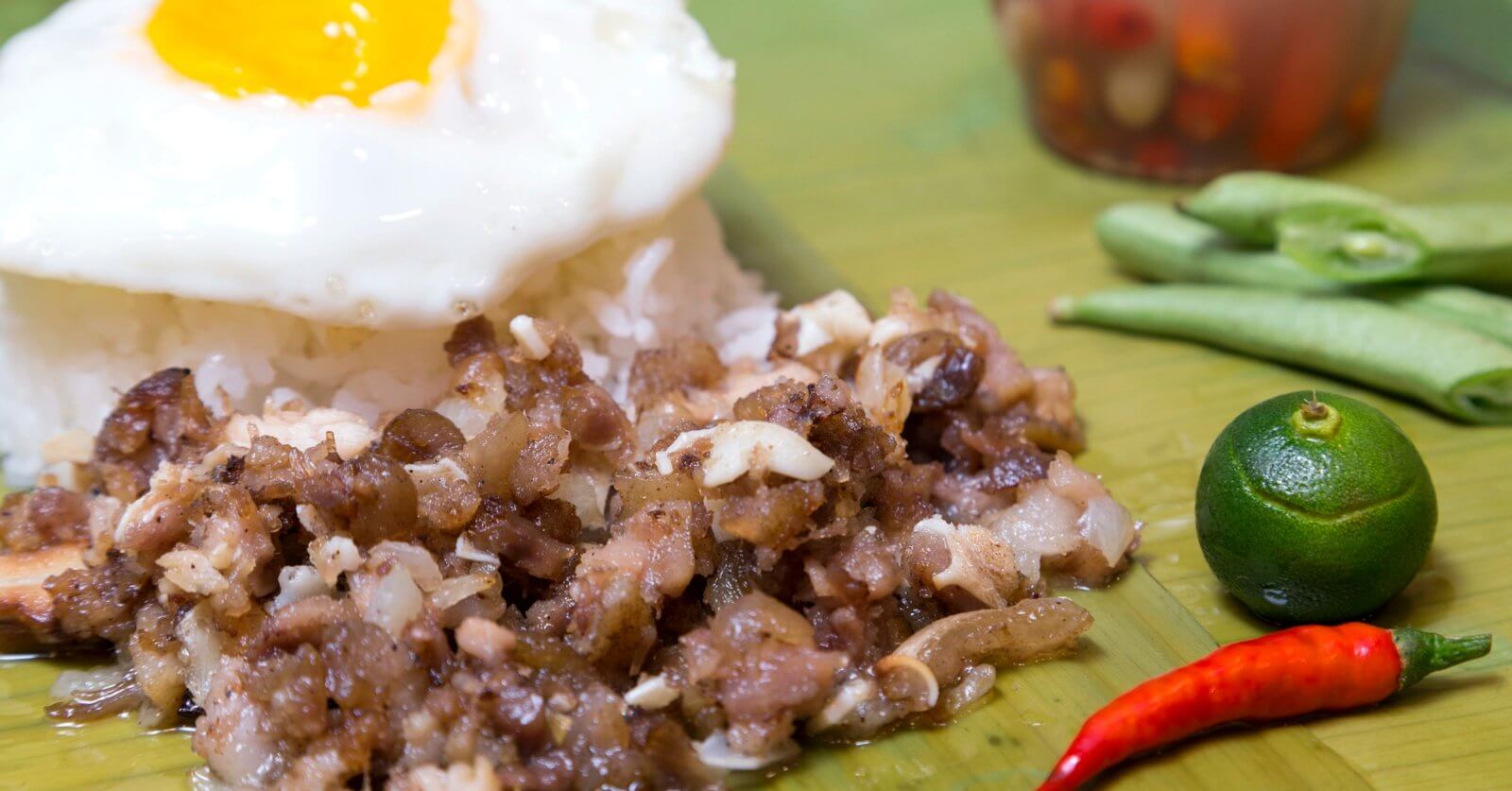 A close-up of traditional Filipino food featuring a serving of garlic rice topped with a sunny-side-up egg. Adjacent to the rice is a portion of sizzling chopped meat, likely pork. Garnishing the plate are a whole calamansi, a red chili pepper, and a few green beans. A condiment jar is blurred in the background.