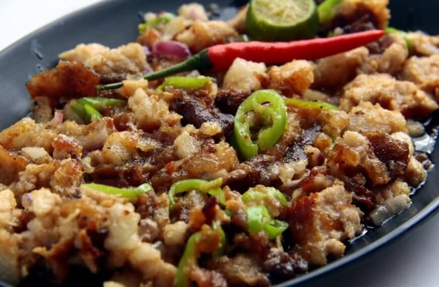 A black oval plate holds a serving of sisig, a popular Filipino dish. This Food Lover's Guide favorite from Laguna consists of chopped pork, onions, and green chili slices topped with a whole red chili. A sliced calamansi lime and crispy bits of pork adorn the top, with visible oil glistening on the surface.