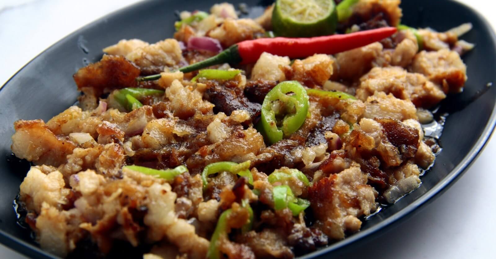 A black oval plate holds a serving of sisig, a popular Filipino dish. This Food Lover's Guide favorite from Laguna consists of chopped pork, onions, and green chili slices topped with a whole red chili. A sliced calamansi lime and crispy bits of pork adorn the top, with visible oil glistening on the surface.