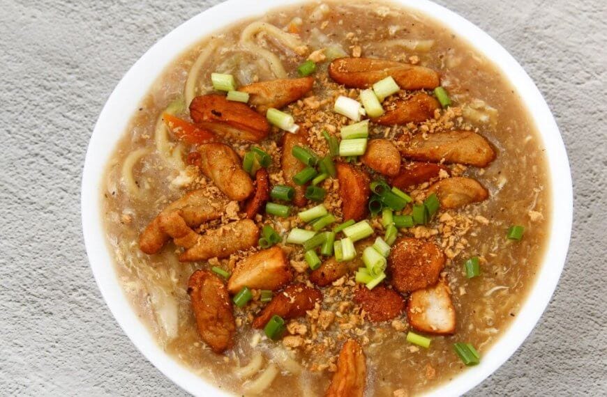 A top-down view of a hearty bowl of noodle soup garnished with chunks of fried chicken, sliced green onions, and bits of crunchy fried garlic. The soup's rich broth is visible, with noodles submerged beneath the toppings. This delectable dish could rival the best Lomi places in Batangas. The bowl rests on a textured gray surface.