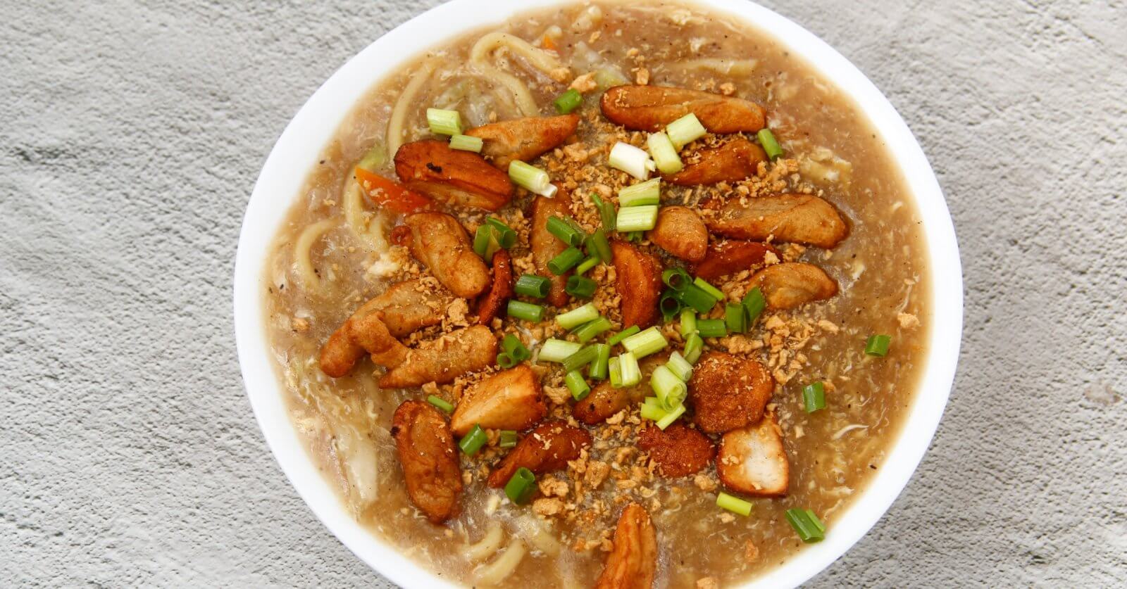 A top-down view of a hearty bowl of noodle soup garnished with chunks of fried chicken, sliced green onions, and bits of crunchy fried garlic. The soup's rich broth is visible, with noodles submerged beneath the toppings. This delectable dish could rival the best Lomi places in Batangas. The bowl rests on a textured gray surface.