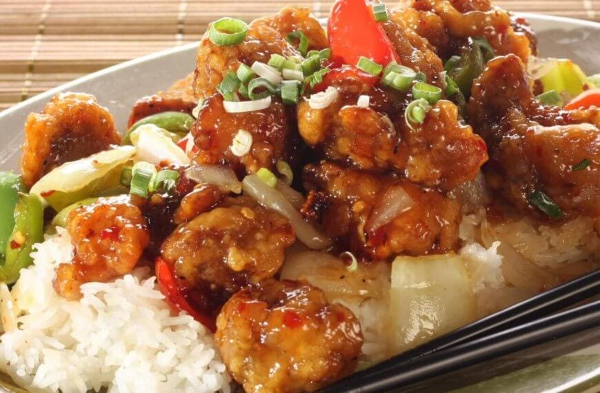 A plate filled with sweet and sour chicken served over white rice is garnished with chopped green onions. The dish includes chunks of chicken coated in a glossy sauce with red and green bell peppers and onions. Black chopsticks rest on the side of the plate, which sits on a wooden surface at Lucky Chinatown.